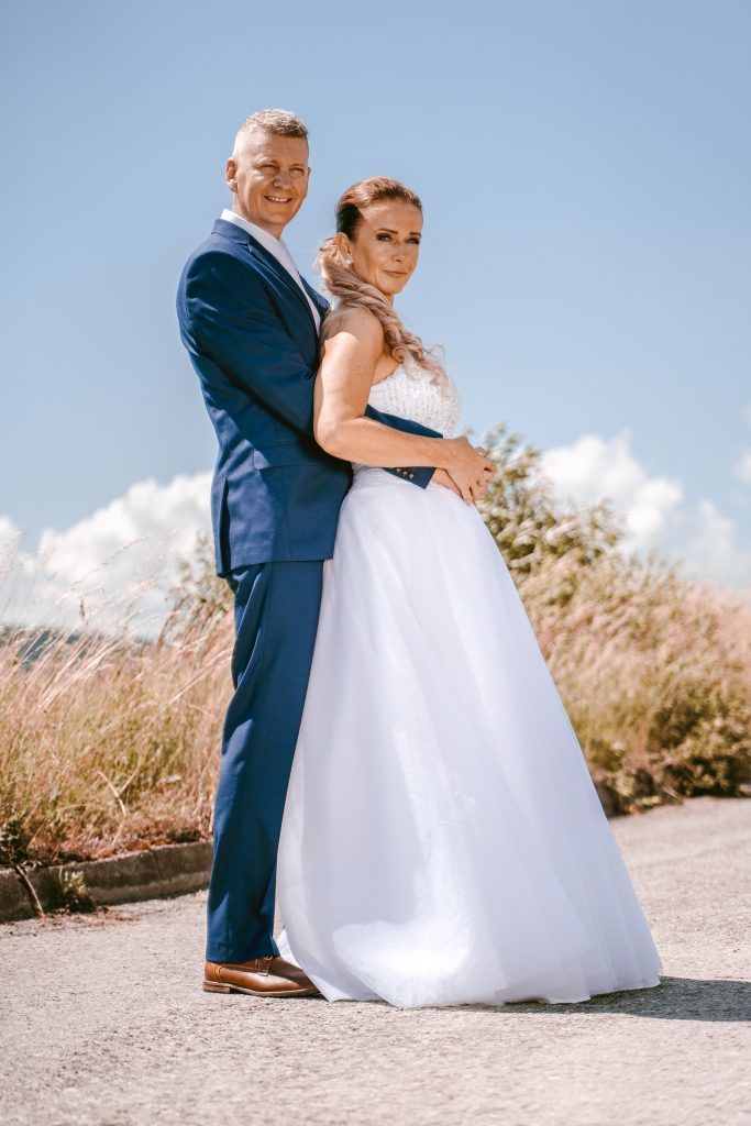 Nevesta objíma ženícha zozadu na poľnej ceste pod letnou oblohou