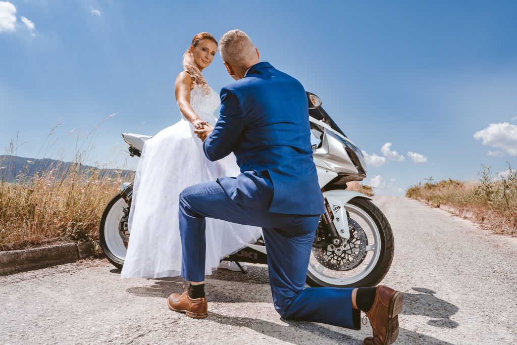Ženích kľačí pred nevestou sediacou na motorke uprostred cesty
