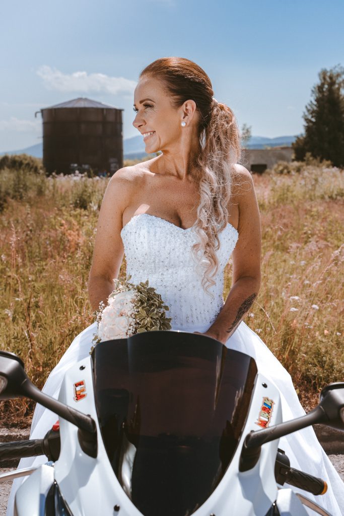 Nevesta sedí na bielej motorke s kyticou, v pozadí letná lúka