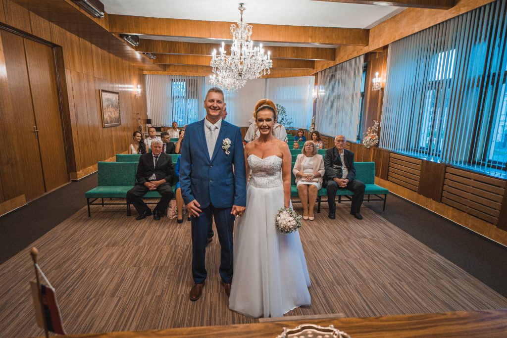 Novomanželia stoja v sobášnej sieni s lustrom, za nimi sedia hostia