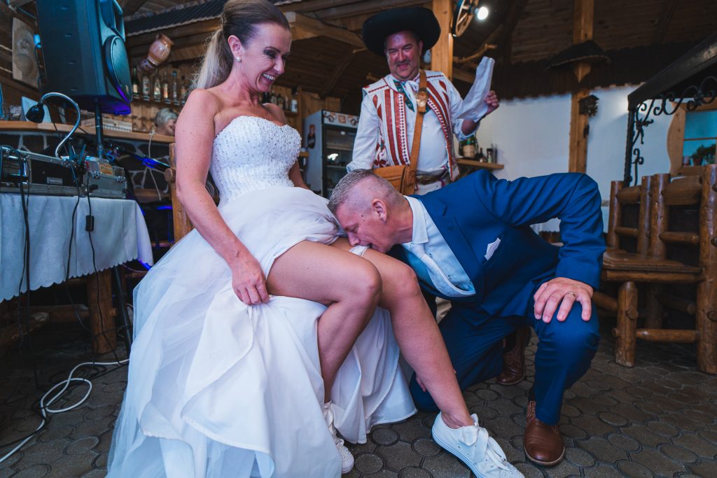 Miro sníma Elle podväzok zubami, v pozadí muž v ľudovom kroji