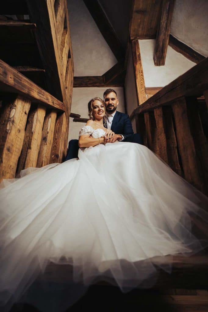 Novomanželia sedia na drevených schodoch, šaty nevesty zaberajú popredie