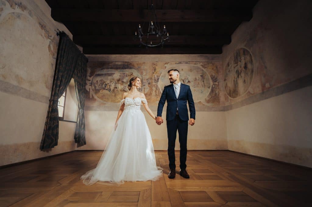 Novomanželia sa držia za ruky v stredovekej sále s freskami na stenách