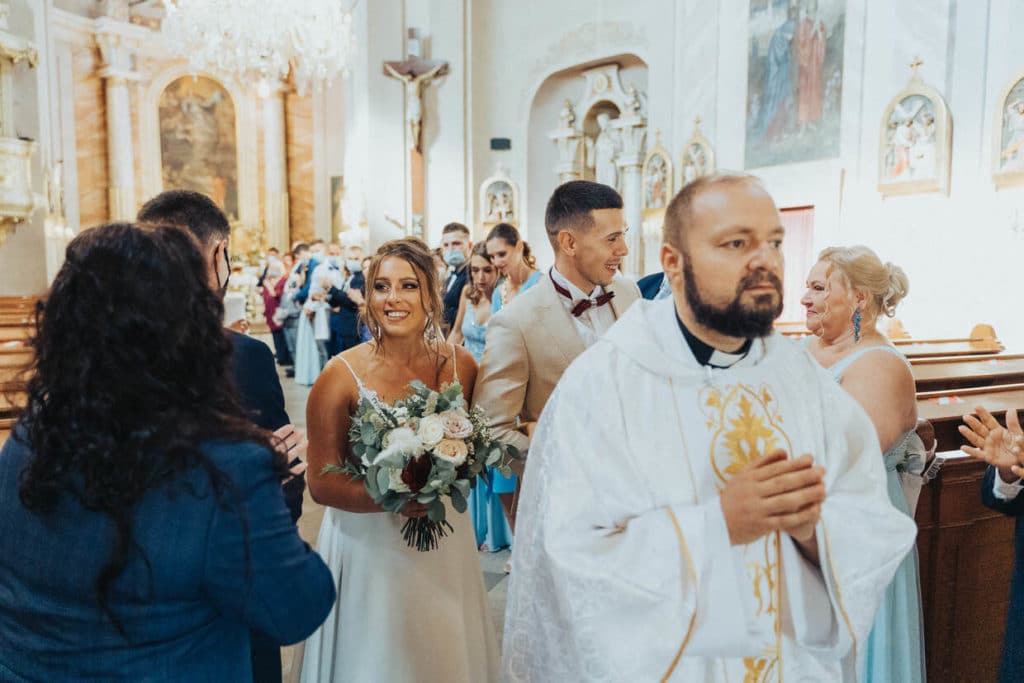 Kňaz v bielom rúchu, za ním nevesta s kyticou a ženích v kostole
