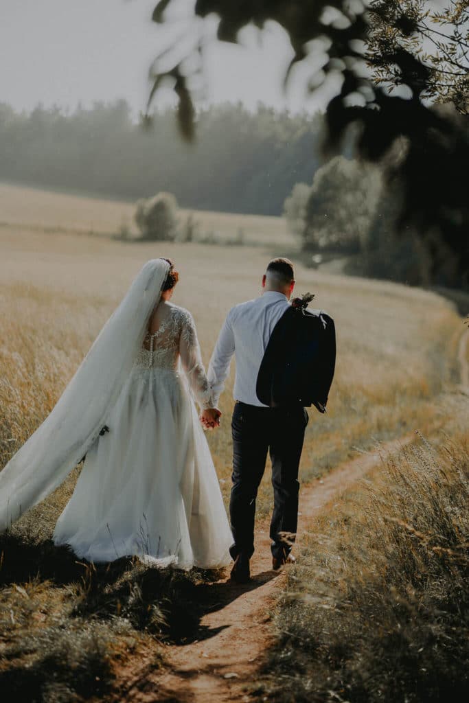 Andrea a Peter kráčajú chodníčkom cez lúku, závoj viaje vo vetre