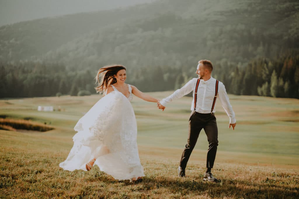Nevesta a ženích bežia ruka v ruke cez lúku, šaty sa vlnia