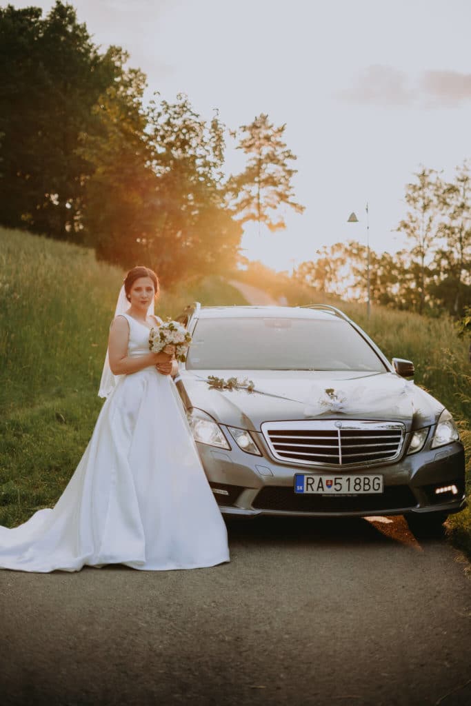 Nevesta s kyticou stojí pred ozdobeným svadobným mercedesom v zapadajúcom slnku