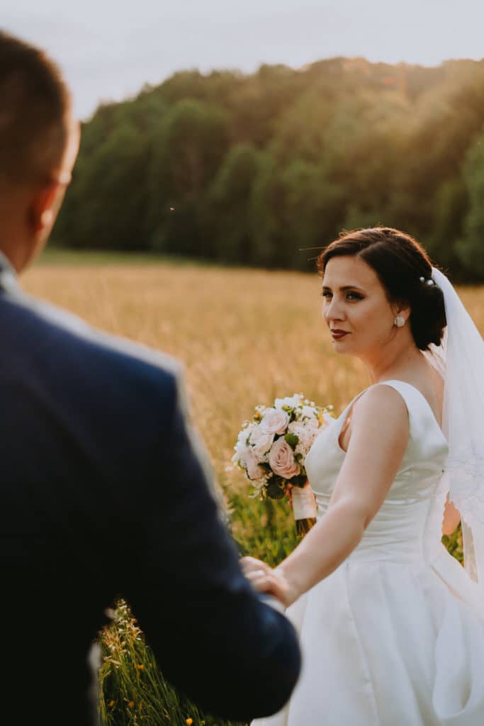 Nevesta so závojom sa ohliada na ženícha, ktorý ju vedie za ruku poľom