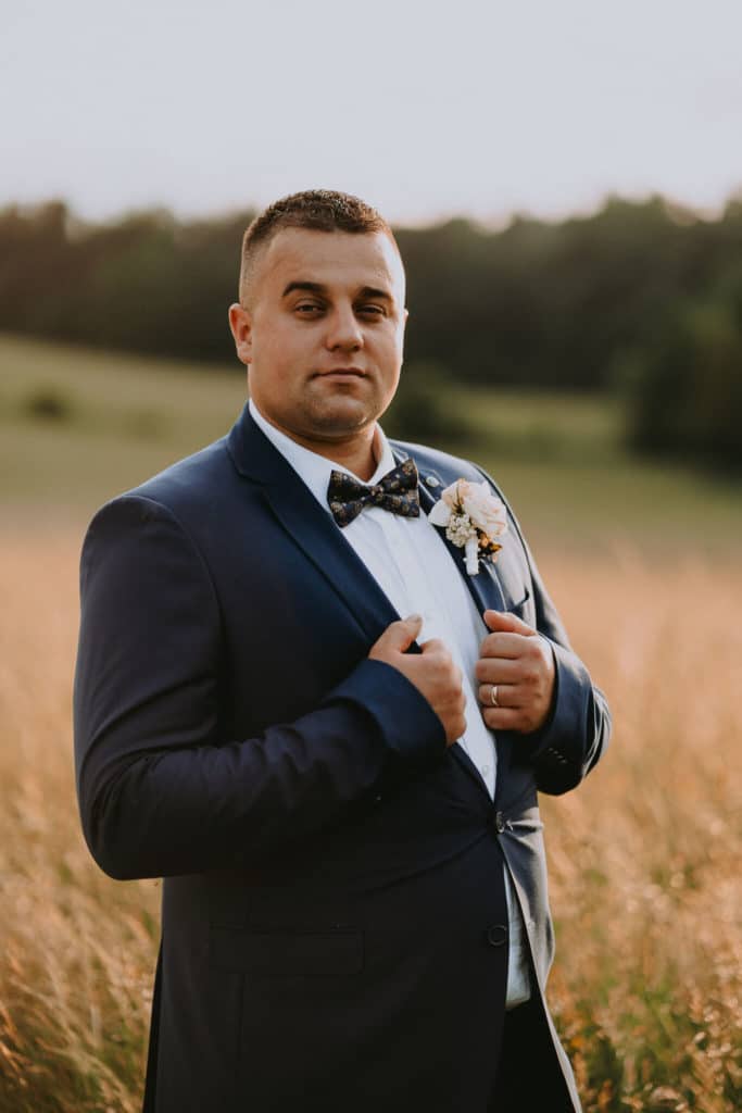 Ženích v tmavomodrom obleku s motýlikom si upravuje sako v poli