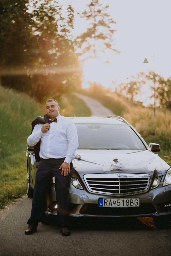 Ženích v bielej košeli sa opiera o kapotu svadobného mercedesu
