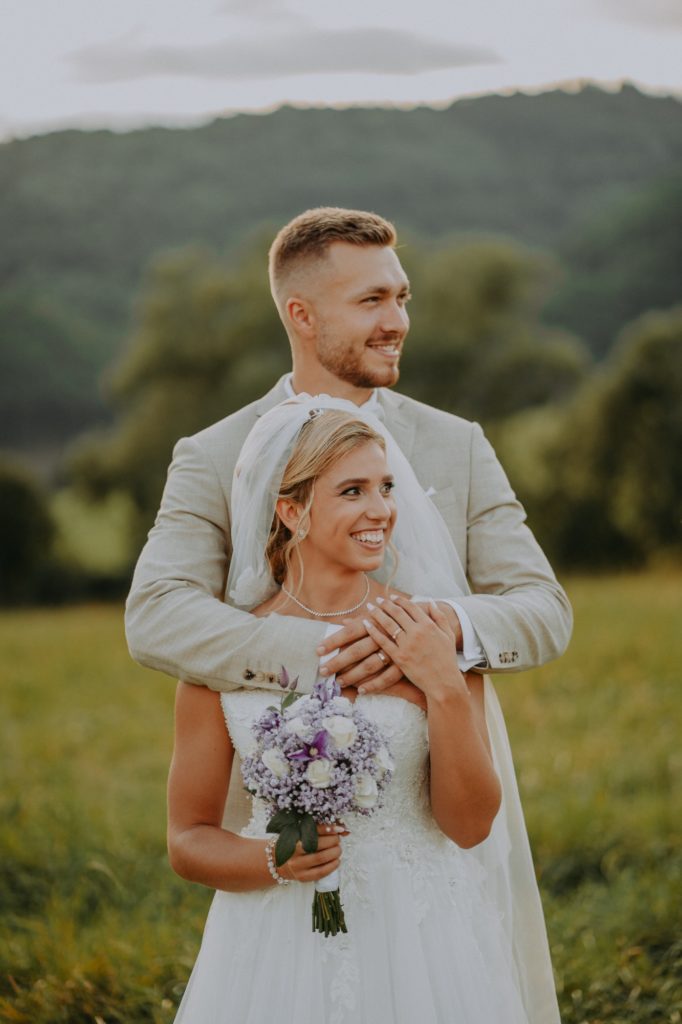 Ženích v béžovom obleku objíma usmiatu nevestu s fialovou kyticou