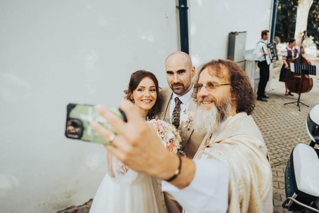 Usmiaty kňaz s bradou si robí selfie s novomanželmi pred kostolom
