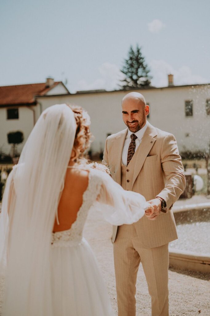 Ženích sa s úsmevom otáča k neveste v šatách s holým chrbtom a dlhým závojom