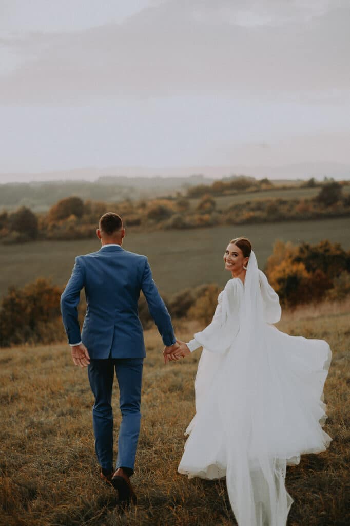 Ženích vedie nevestu za ruku cez lúku, jej šaty sa vlnia vo vetre
