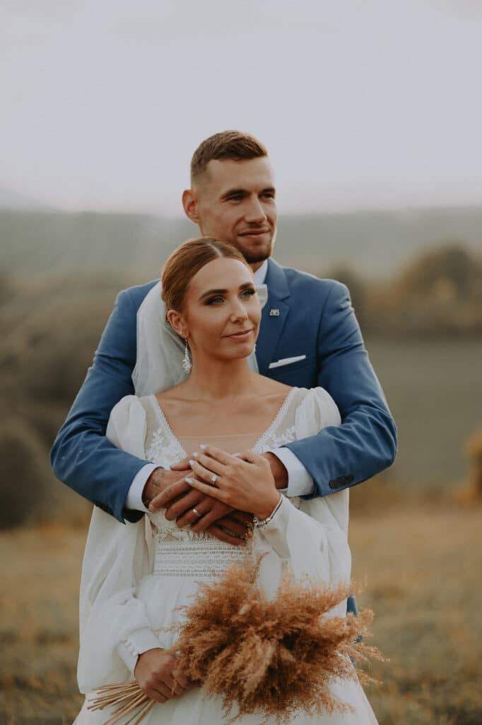 Ženích objíma nevestu zozadu, ona drží kyticu zo sušených tráv