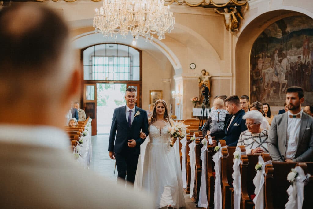 Nevesta kráča uličkou kostola v sprievode muža, ženích ju čaká v popredí
