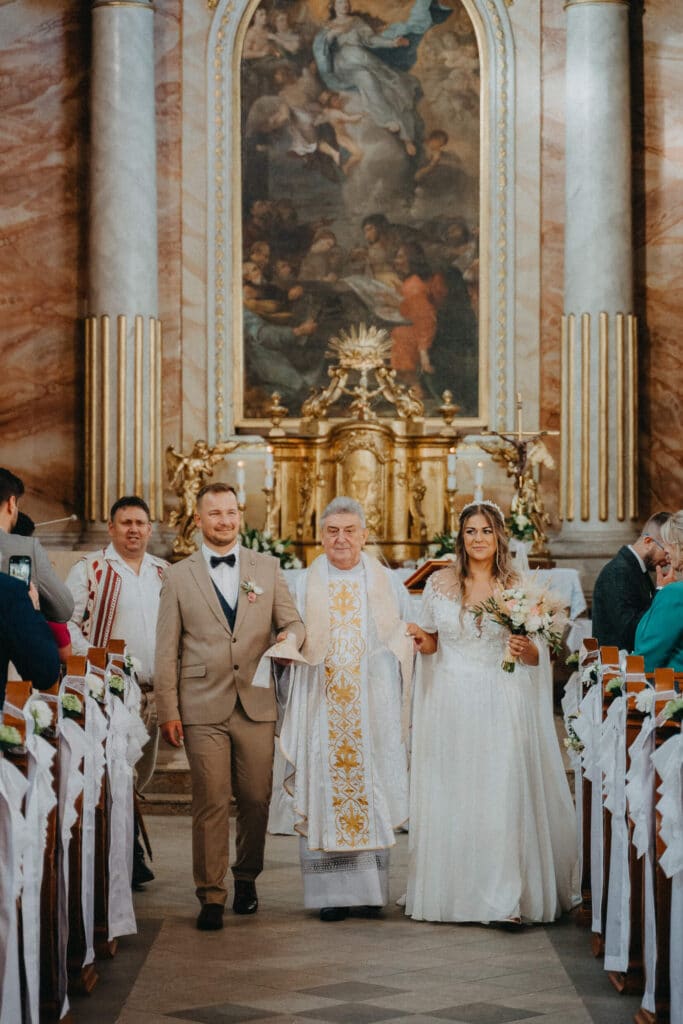 Novomanželia odchádzajú s kňazom uličkou zdobeného barokového kostola