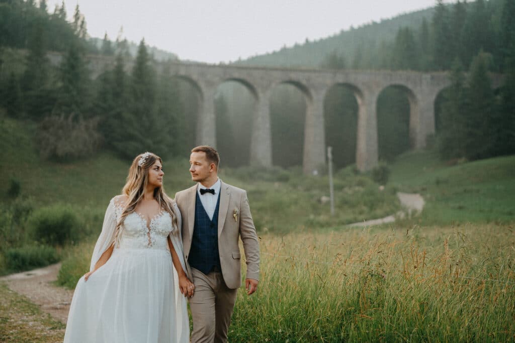 Katka a Michal kráčajú lúkou pred kamenným viaduktom v horách